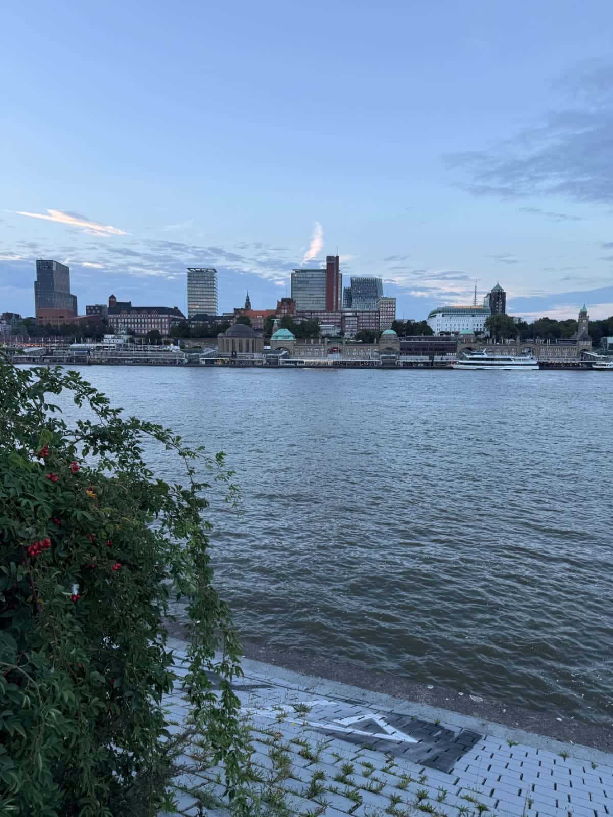 Vista del Elba hacia el perfil urbano de Hamburgo con luz de atardecer, y paseo ribereño y agua en primer plano