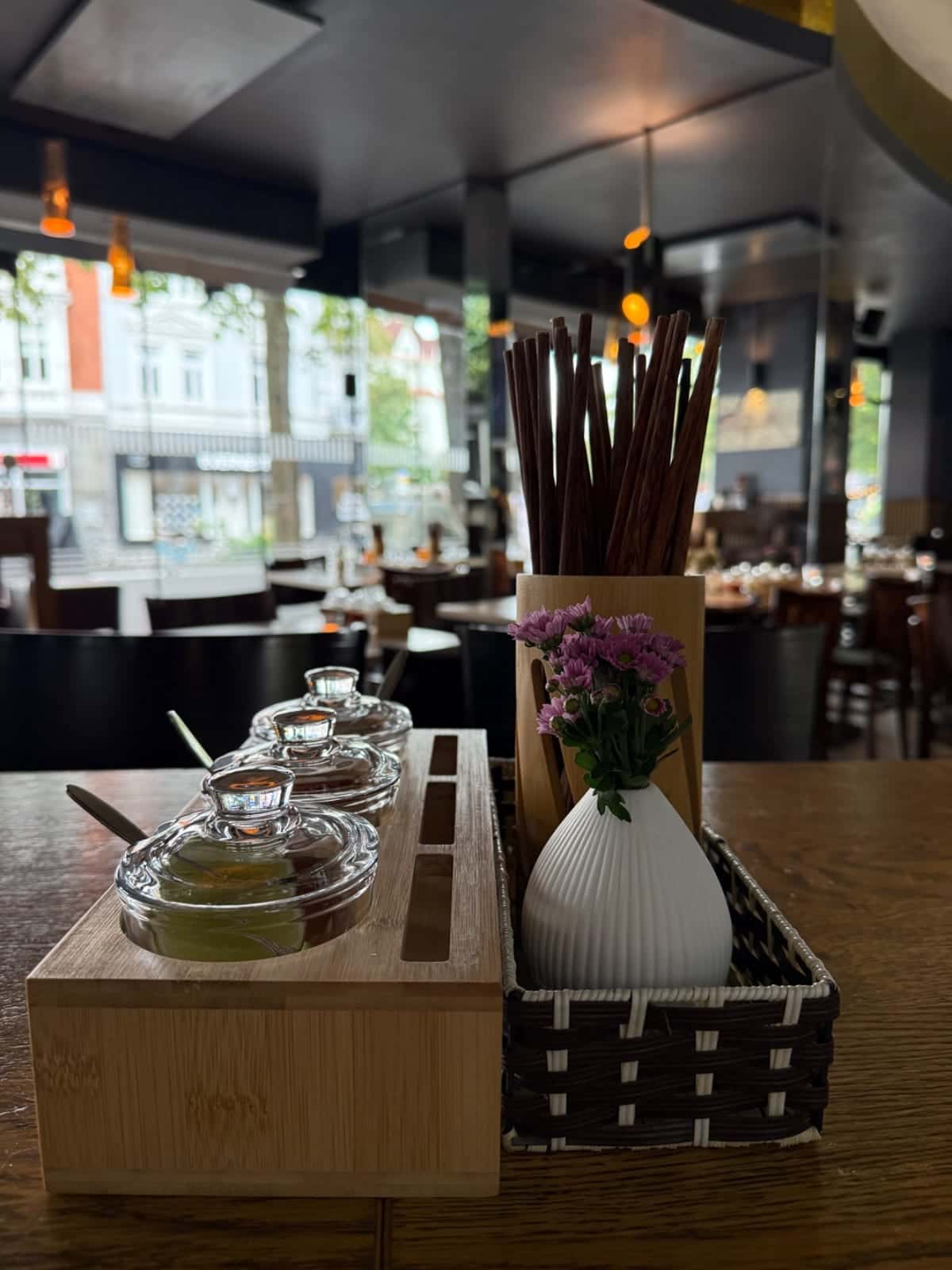 Mesa con frascos de especias, palillos y un pequeño jarrón de flores en Phở Dàm; mesas y grandes ventanales al fondo