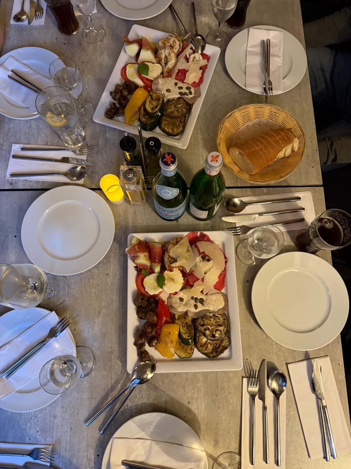 Dos mesas preparadas con grandes bandejas de antipasti, pan, vasos y botellas