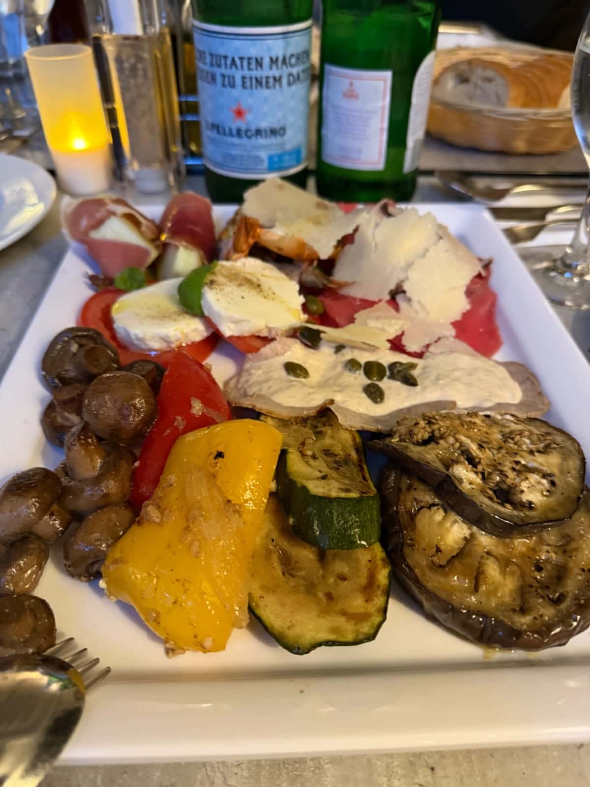 Plato de antipasti con verduras a la parrilla, aceitunas, queso y jamón sobre una mesa con botellas de bebida y una vela