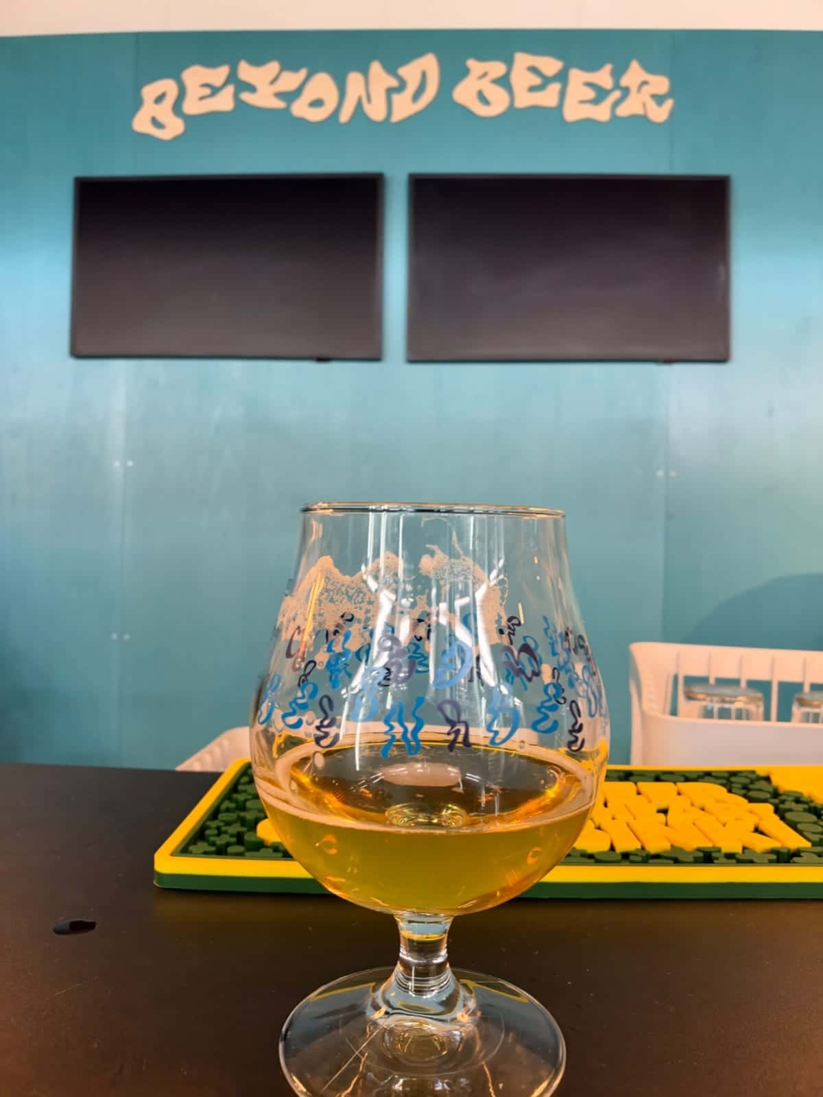 Una copa de cerveza con una bebida clara está sobre una mesa frente a una pared turquesa con el letrero Beyond Beer