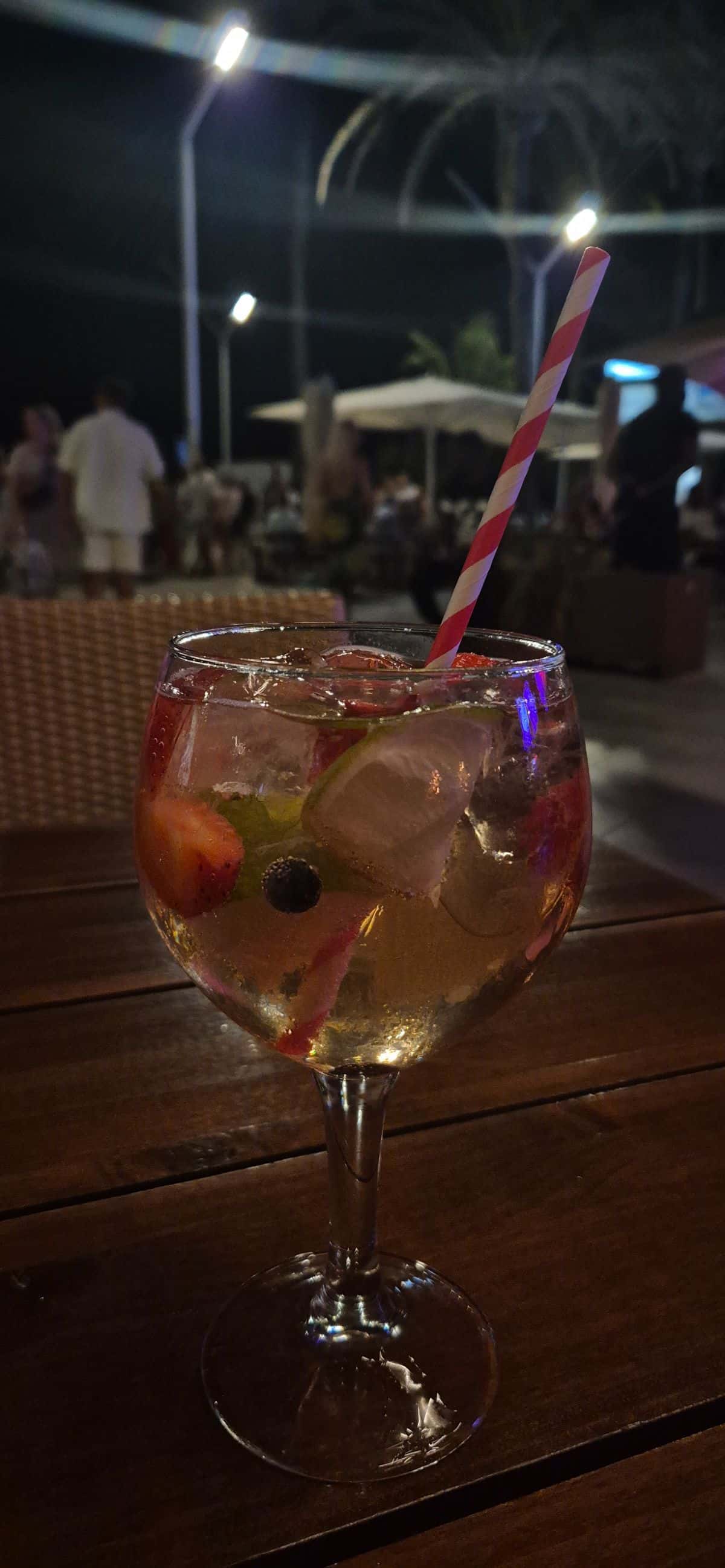 Cocktail im Glas mit Eis, Früchten und rot-weißem Strohhalm auf einem Holztisch, im Hintergrund Gäste im Freien bei Nacht