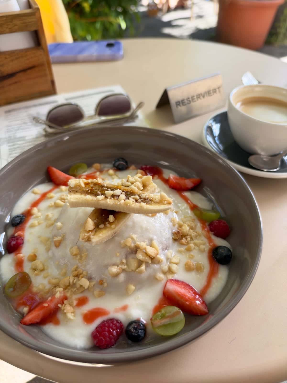 Schüssel mit Joghurt, Beeren, Nüssen und Obst auf einem Tisch, daneben eine Tasse Kaffee und eine Reserviert-Karte