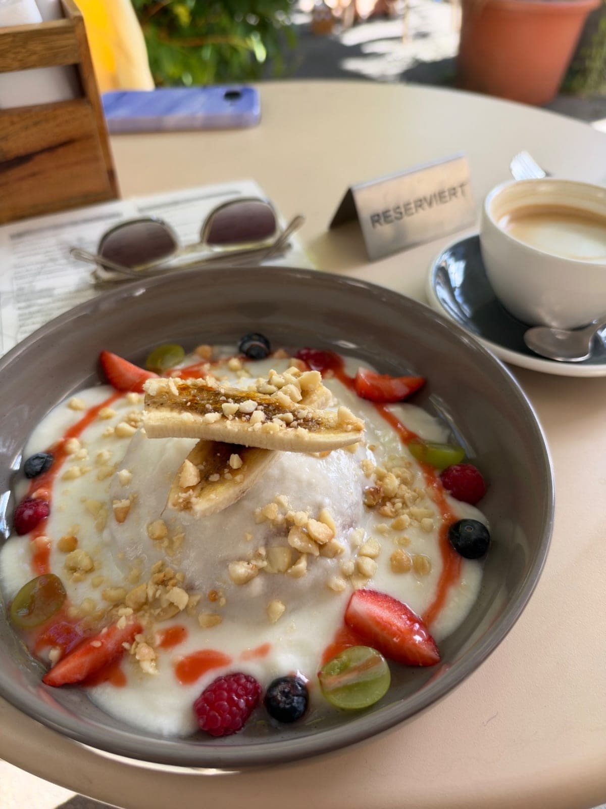 Schüssel mit Joghurt, Beeren, Nüssen und Obst auf einem Tisch, daneben eine Tasse Kaffee und eine Reserviert-Karte